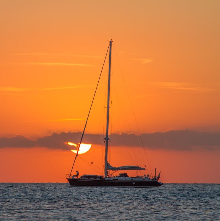 Modern sailboat silhouetted against an orange sunset on calm ocean water